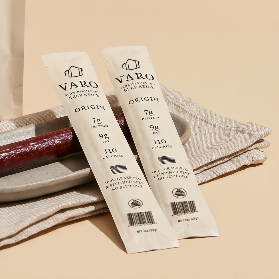 Two wrapped ORIGIN VARO slow-fermented beef sticks resting on a plate and linen beside an unwrapped stick